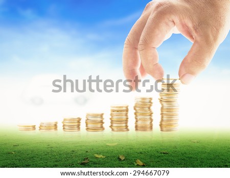 Human hand adding a golden coin in the final row of golden coins over blurred white car on beautiful blue sky background. Concept for money coin, insurance, buying, renting, repair, fuel, service.