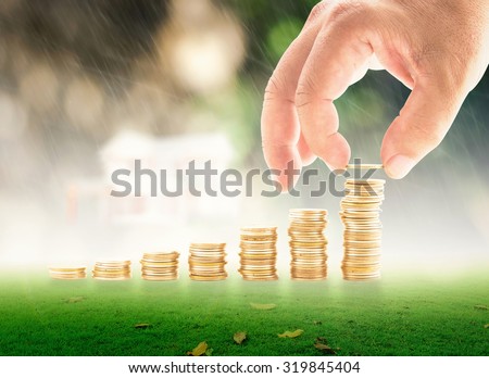 Human hand adding a golden coin in the final row of golden coins over blurred bokeh beautiful house on rainy with nature background. Concept for money coin, insurance, buying, renting, service.