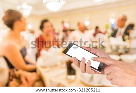male hand is holding a modern touch screen phone and blur image of wedding party  in large hall for background usage.