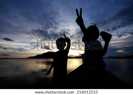 Silhouetted kids (sibling, Excited and joyous sister showing peace hand signal while drinking an energy drink and her younger brother who looks not so happy, also with peace hand signal) during sunset