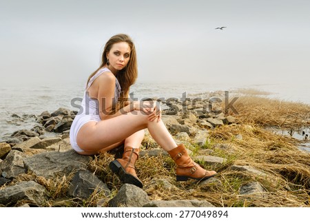 Beautiful Girl sits on stone beach. Young woman sits in sunny day on the river bank. Long Shot. LS