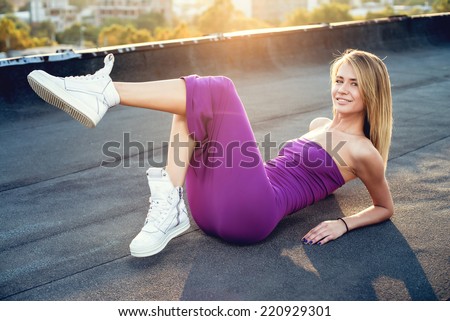 Beautiful young woman wearing a purple dress with a pair of white sneakers on the roof. Sunset