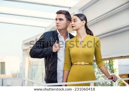 Man and woman wearing classic clothes, him in white shirt and leather jacket, her in yellow dress standing together with the business center on the background. Outdoor fashion shot.