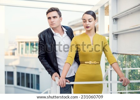 Man and woman wearing classic clothes, him in white shirt and leather jacket, her in yellow dress standing together with the business center on the background. Outdoor fashion shot.