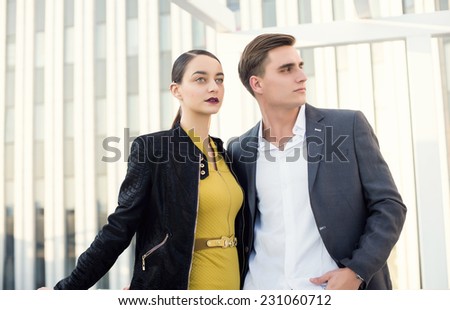 Man and woman wearing classic clothes, him in white shirt and grey jacket, her in yellow dress with leather jacket standing together with the business center on the background.