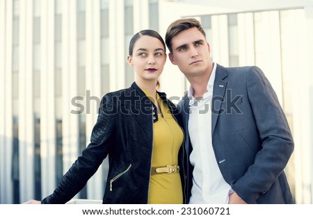 Man and woman wearing classic clothes, him in white shirt and grey jacket, her in yellow dress with leather jacket standing together with the business center on the background.