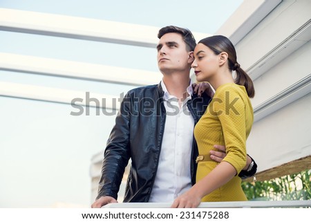 Man and woman wearing classic clothes, him in white shirt and leather jacket, her in yellow dress jacket standing together with the business center on the background. Outdoor fashion shot.