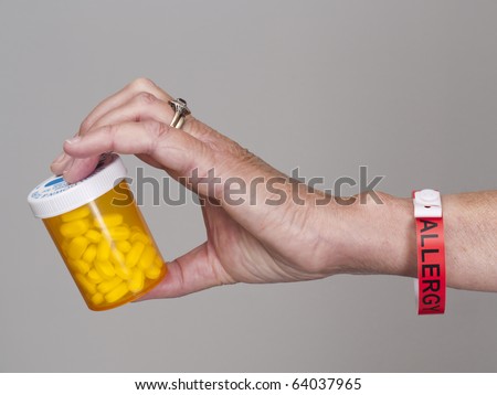 Hand with red allergy band holding container of pills to treat allergies. Gray background.