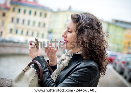 the girl does makeup, cute girl paints her lips look in the mirror in the center of a city, on the embankment