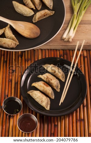 Freshly cooked Chinese style pot stickers with soy sauce, pepper sauce and raw green onion. Four pot stickers are plated beside the cooking pan.