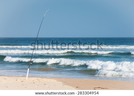 Fishing rod on empty beach with two surfers in the ocean at Lake Conjola, Australia in the late afternoon