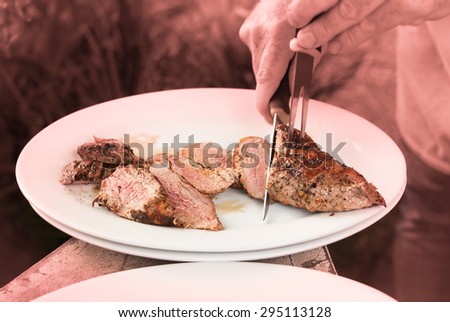 Cutting of juicy medium rare to well roasted pork meat - sirloin steak - on white plates in the garden barbecue party - burgundy red tinted photography background