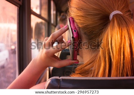 Attractive teenage girl with red orange hair talking over her pink smartphone in public transport looking through window in a sunsetting light - rear view