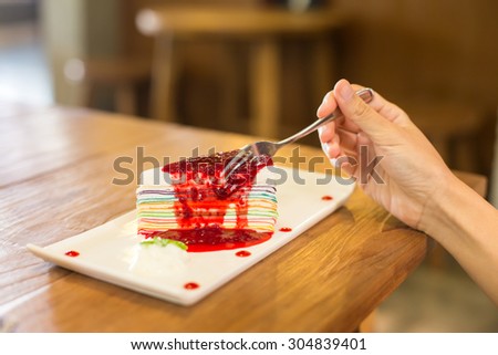 Rainbow crape cake on white plate with woman\'s hand going to eat.