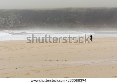 SENNEN COVE, CORNWALL, ENGLAND - OCTOBER 24: Surfer walking across beach to get to water on cold, wet, misty, miserable British autumn day, shown on 24 October 2014 in Sennen Cove