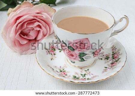 Tea in a china cup and saucer, with pink rose pattern and pink cut rose. On a white, painted, distressed table top.