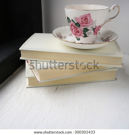 Cup of tea in a vintage china cup and saucer on top of a stack of hardback books on a windowsill. Square photo.