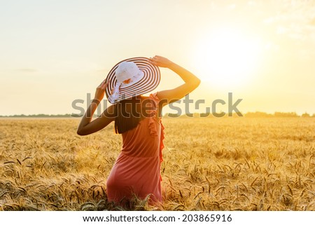 Beautiful young woman with brown hear wearing rose dress and hat with raised arms enjoying outdoors looking to the sun on perfect wheat field on sunset