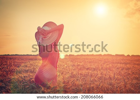 Beautiful young woman with brown hear wearing rose dress and hat with raised arms enjoying outdoors looking to the sun on perfect wheat field on sunset