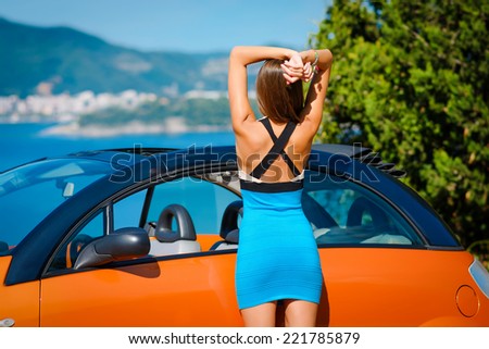 Beautiful young woman with long hair standing back near orange cabriolet at the Mediterranean sea coast