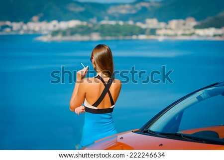 Beautiful young woman with long hair smoking and standing near orange cabriolet at the Mediterranean sea coast