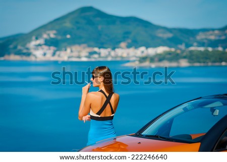 Beautiful young woman with long hair smoking and standing near orange cabriolet at the Mediterranean sea coast