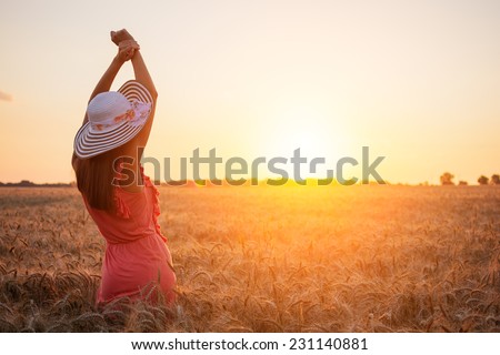 Beautiful young woman with brown hear wearing rose dress and hat enjoying outdoors looking to the sun on perfect wheat field on sunset
