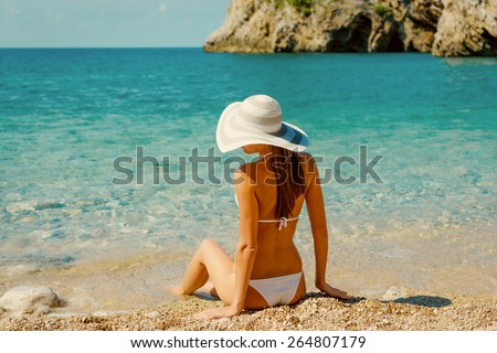 Sexy back of beautiful woman in white bikini with creative white sunglasses on Mediterranean sea coast. Montenegro, South Europe. Retro vintage toned image, film simulation.