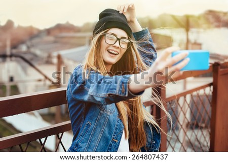 Young sexy blonde hipster woman posing for selfie and laughing. Wearing jeans jacket, hipster black hat and glasses. Lifestyle portrait bright with sun shine.