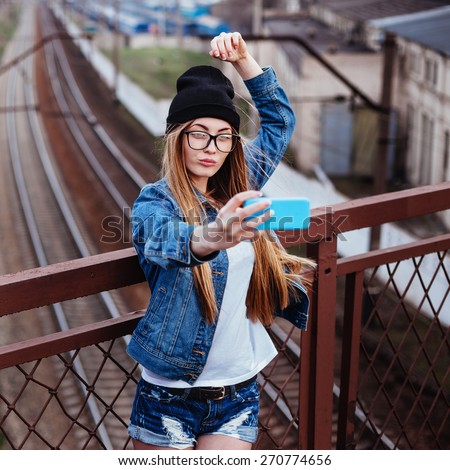 Outdoor lifestyle portrait of young sexy blonde hipster woman posing for selfie and laughing. Wearing jeans jacket, hipster black hat and glasses.