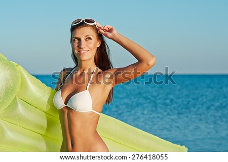 Beautiful happy woman in white bikini and creative white sunglasses with yellow inflatable mattress on the beach.