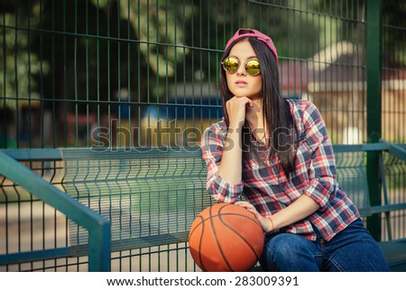 Outdoor lifestyle portrait of pretty young girl, wearing in hipster swag grunge urban style and creative sunglasses. Basketball court and orange ball. Retro vintage toned image, film simulation.