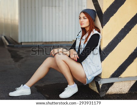 Outdoor lifestyle portrait of pretty young sitting girl, wearing in hipster swag grunge style urban background. Red hair