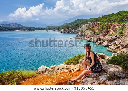 Portrait of beautiful tanned woman sitting at the sea coast. Hot summer day and bright sunny light. Panoramic view on sea shore near Kemer, Antalya, Turkey.