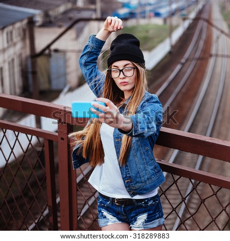 Outdoor lifestyle portrait of young sexy blonde hipster woman posing for selfie and laughing. Wearing jeans jacket, hipster black hat and glasses.
