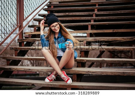 Young sexy blonde hipster woman posing for selfie and laughing. Wearing jeans jacket, hipster black hat and glasses. Lifestyle portrait
