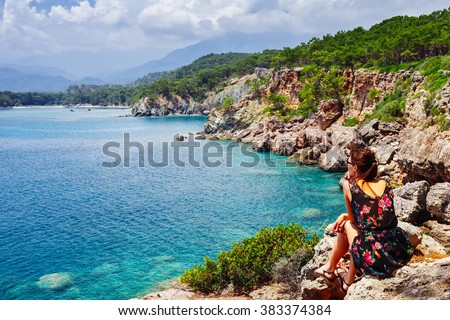 Portrait of beautiful tanned woman sitting at the sea coast. Hot summer day and bright sunny light. Panoramic view on sea shore near Kemer, Antalya, Turkey.