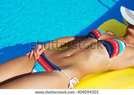 Portrait of beautiful tanned woman relaxing in swimming pool in striped swimwear with yellow inflatable mattress. Creative gel polish manicure and pedicure. Hot summer day and bright sunny light.