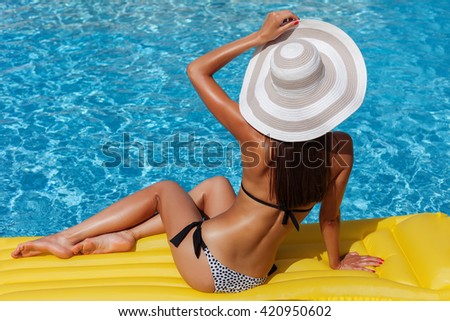 Portrait of beautiful tanned woman relaxing in bikini and hat in swimming pool. Gel polish red manicure. Hot summer day and bright sunny light.