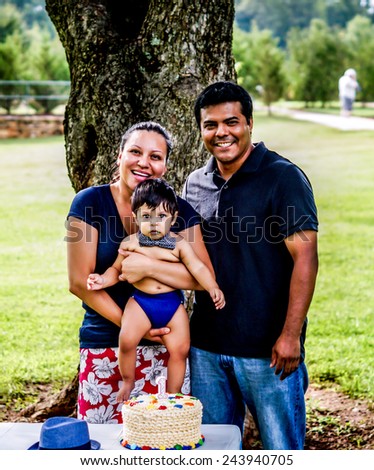 Latino mother, father, and baby with frosting on his face outside by a tree with a birthday cake