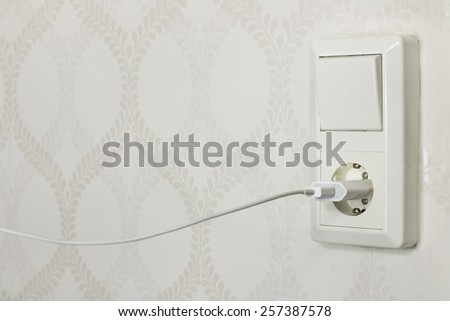 A combined wall outlet and light switch, with a cell phone charger plugged in, and white wall paper background.