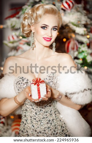 Beauty fashion woman with Christmas gift box, new year tree background. Vogue style sexy girl. Gorgeous girl in luxury fur at Xmas party. Luxury life concept. Retro style girl makeup and jewelry model