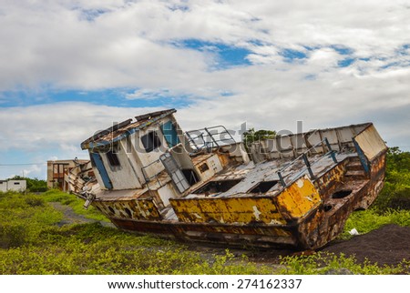 Rusty yellow and blue fishing boat on the shore in Isabela Island, Galapagos, Ecuador. Vessel standing on the grass, sky with white clouds. Vessel abandon on the shore, not needed any more or broken.