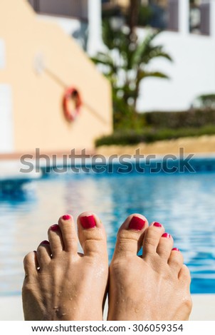 Relaxing by the swimming pool. Woman feet in the foreground and swimming pool in the background. Red nail polish on the toes. Blurry background. Resting, lazy time, summer vacation, summer holidays.