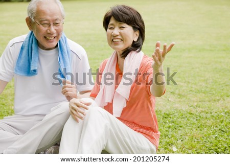 The elderly couple in exercise clothes having a conversation upon sitting on the grass of the park