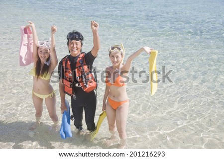 Two swimsuit women taking a commemorative photograph with a webbed foot in the water\'s edge and the man with a diving suit