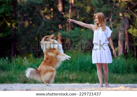 Young beautiful girl in white dress playing with her dog collie on a background of forest. Dog standing on hind legs