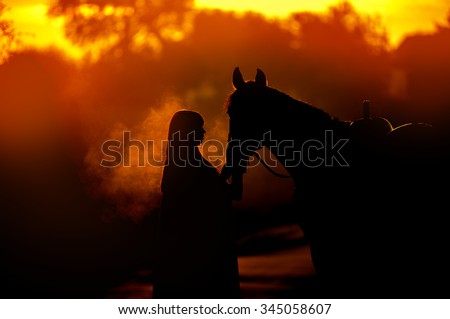 Silhouette of a girl and a horse on a background of dawn. A man standing near a horse that breathes steam.