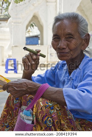 Mingun, Myanmar - February 28, 2014: Old Asiatic woman smoking a cigar outside a temple. The old woman sells fans and cigars to the tourist outside the religious place. She was smiling at me