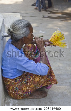 Mingun, Myanmar - February 28, 2014: Old Asiatic woman smoking a cigar outside a temple. The old woman sells fans and cigars to the tourist outside the religious place.